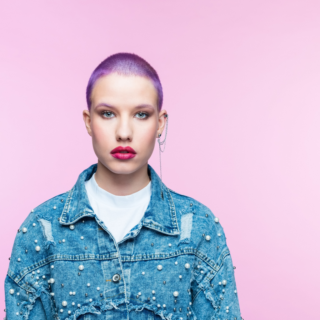 punk rock girl with shaved head wearing silver jewelry on one ear. Red lip stick, purple hair and studded denim jacket pearls with pink background.