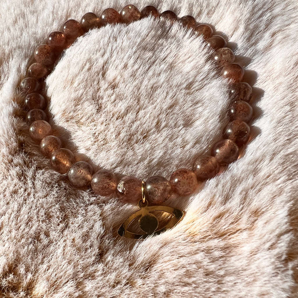 Blush coloured Muscovite bead stretch bracelet with gold eye charm sitting on a pink fluffy pillow in the sunshine. Eye charm meaning intuition.  