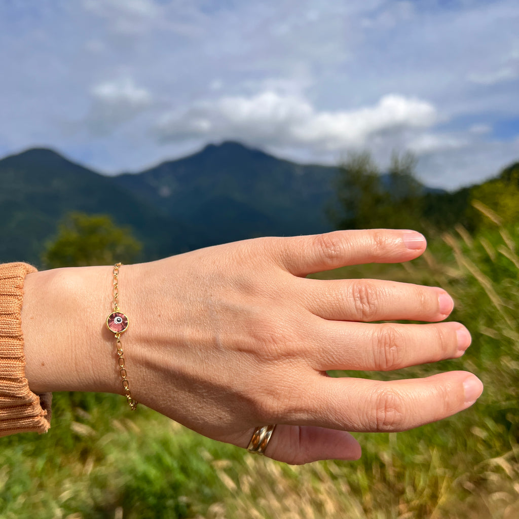 Mountains and green lush meadows back ground,  hand model wearing gold bracelet with rose coloured crystal eye charm and lobster clasp.