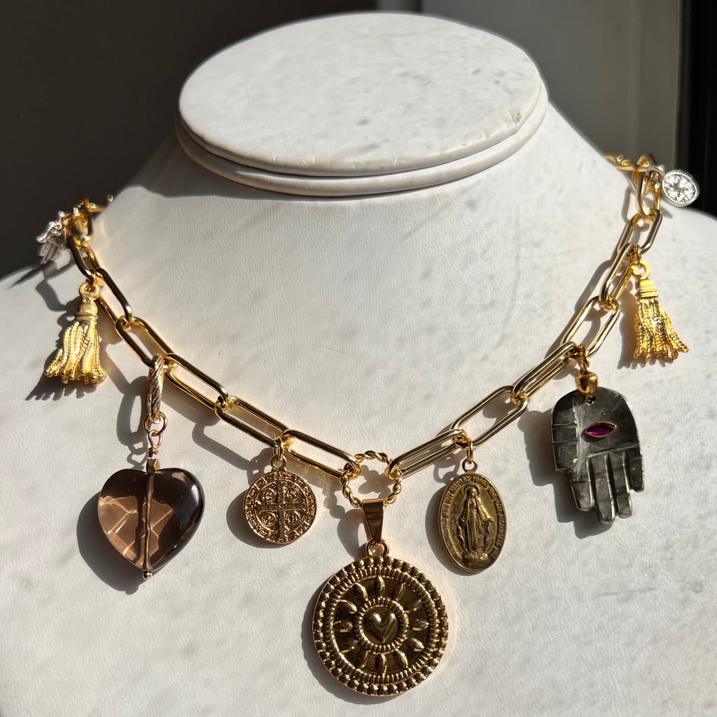 Gold charm bracelet on a mannequin bust with various charms against a neutral background, deity, virgin Mary, hamsa hand. pyrite, heart.
