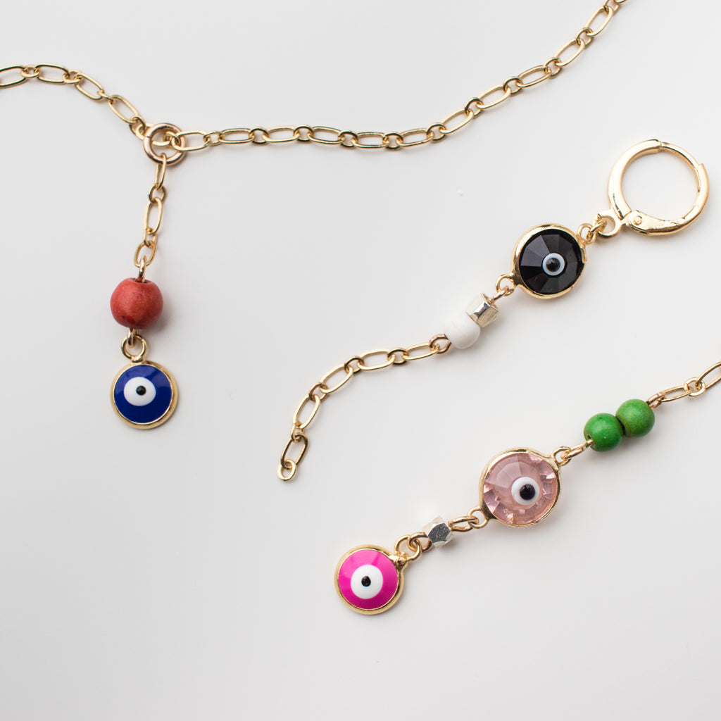 Gold necklace with an enamel cobalt blue eye charm and orange coral bead. Artisan collection with gold mini hoop earrings with light pink, black & hot pink crystal eye charms. Magnesite beads in white and vibrant green, mismatched style earring.